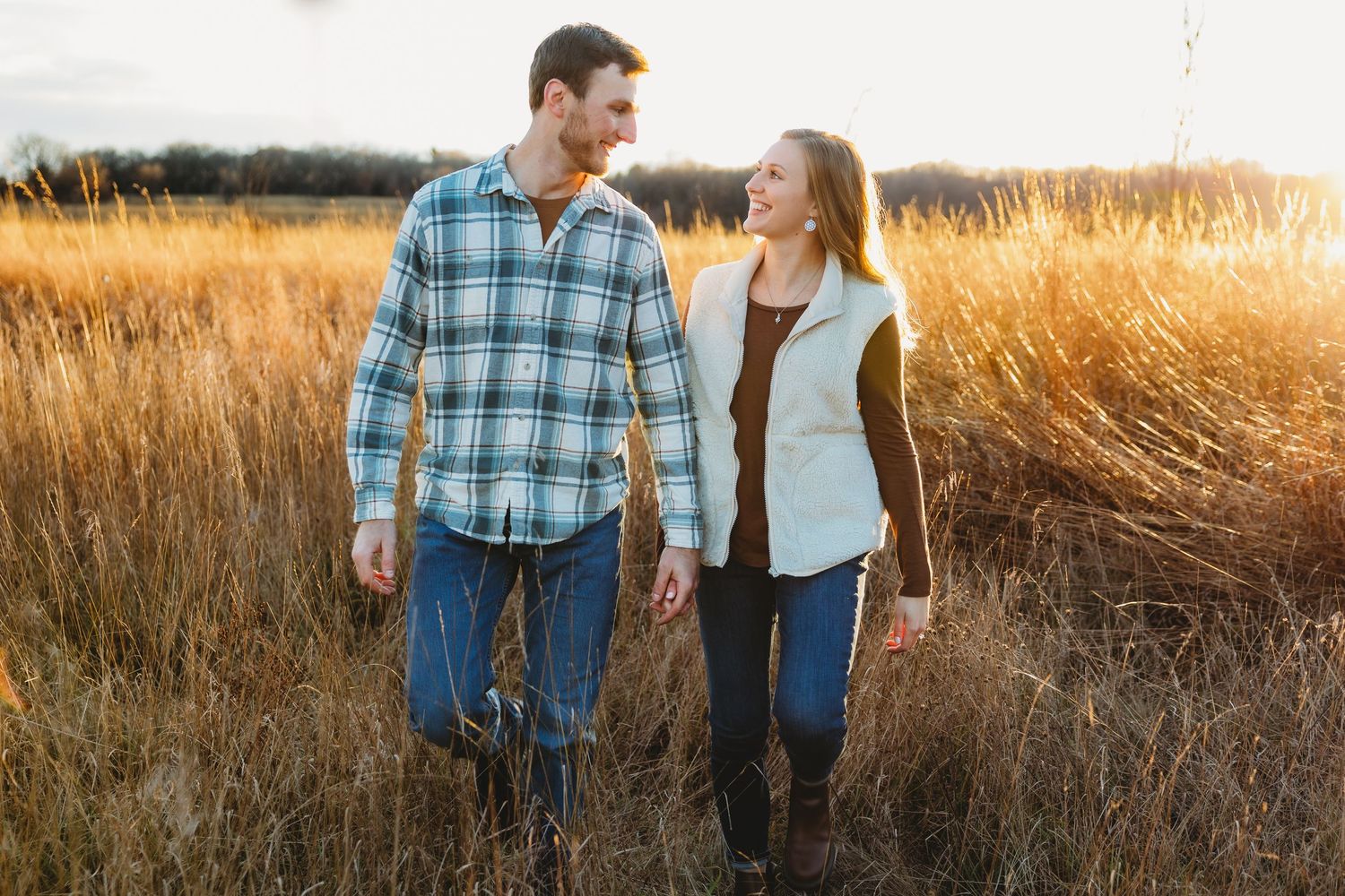 A couple walks together through a golden wheat field at sunset while holding coffee cups.