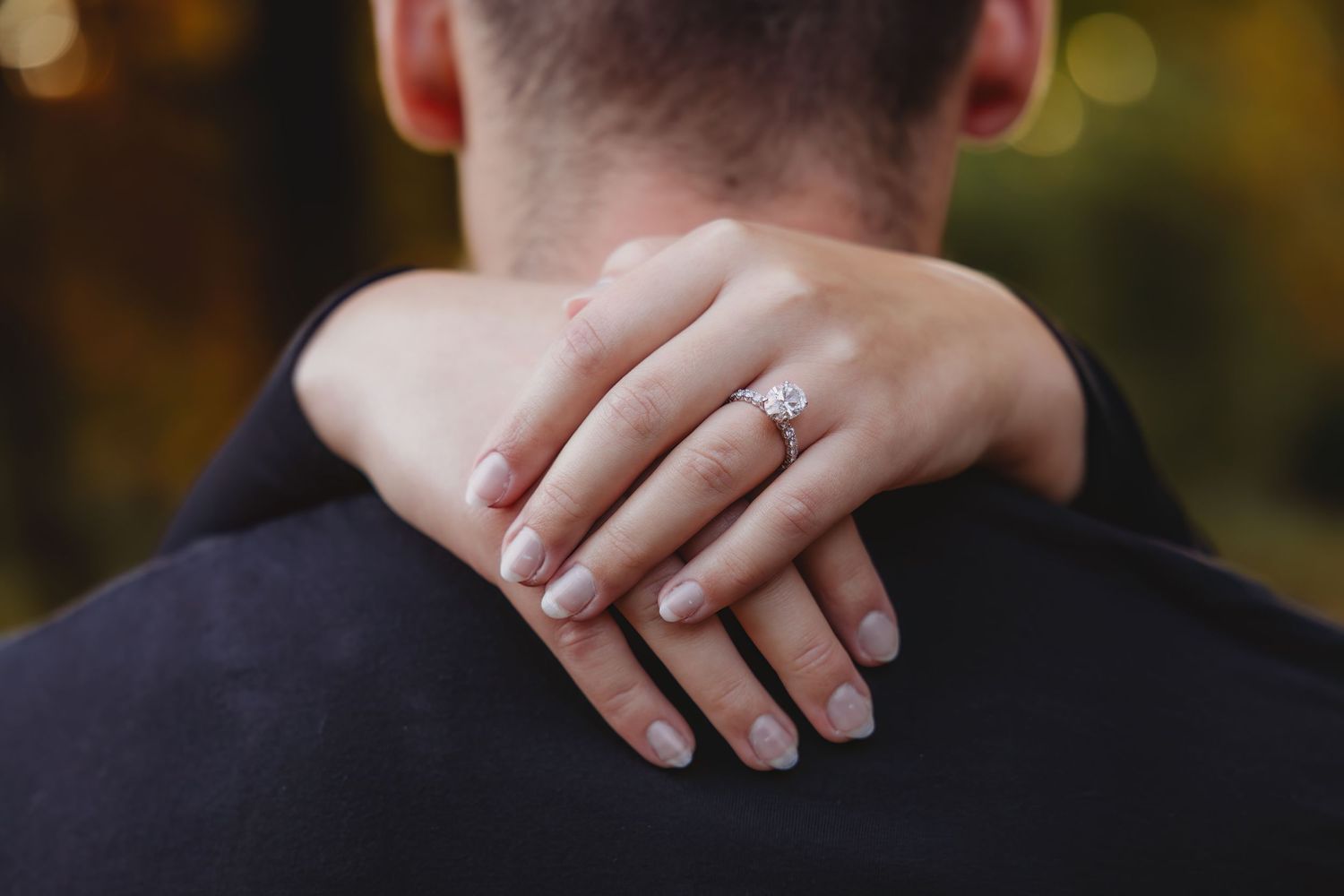 Close-up view of hands showing off a sparkling engagement ring with autumn foliage in the background.