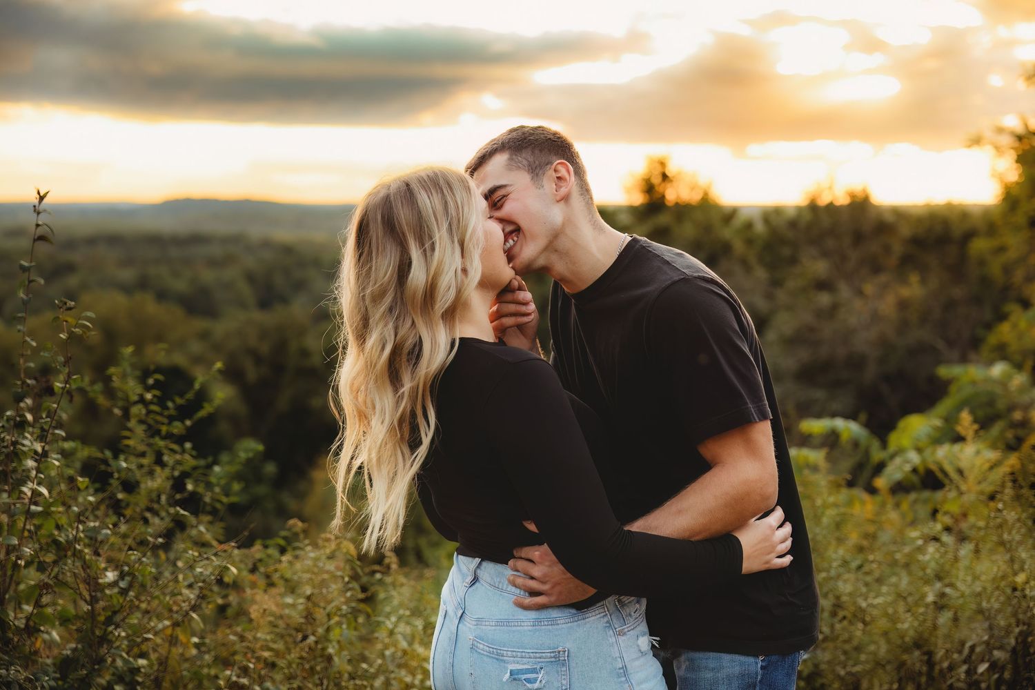 A couple embraces during sunset in a scenic mountain overlook surrounded by lush greenery and golden light.