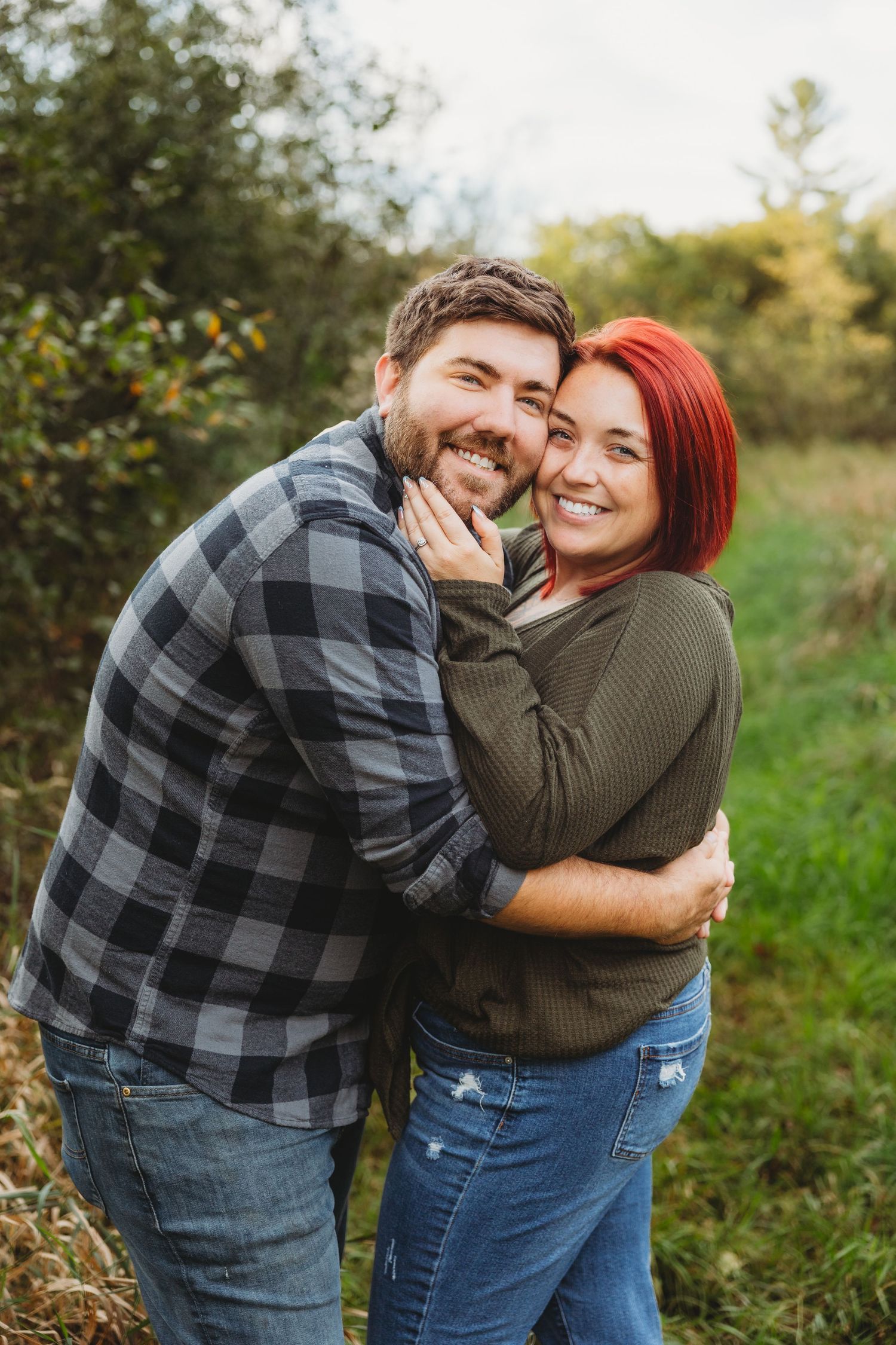 A couple laughs and embraces while posing for photos in a natural outdoor setting with green foliage.