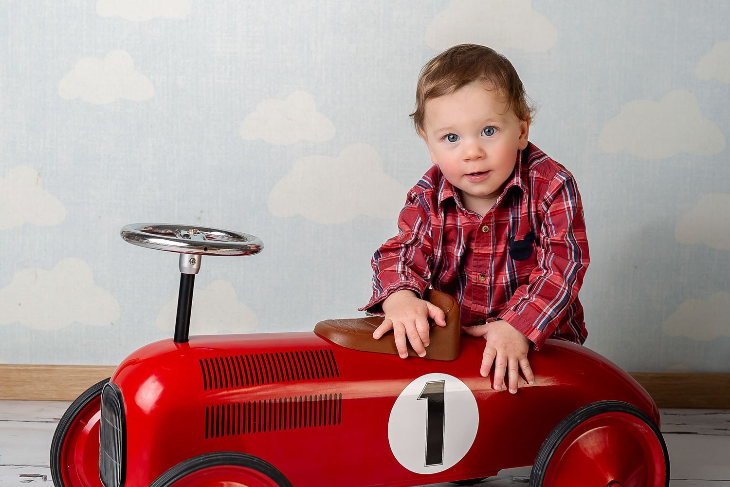 A young child in a red plaid shirt poses with a vintage-style red pedal car toy against a cloudy backdrop.