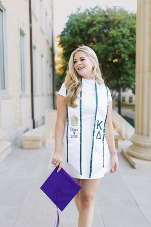 Graduate in white dress holds purple diploma folder while walking through outdoor colonnade.