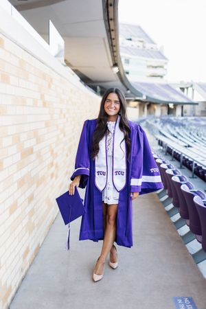 Graduate in purple graduation gown and white dress walks along stadium corridor with diploma in hand.