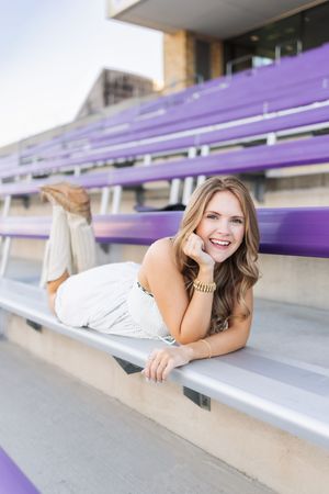 Smiling person lying on purple stadium bleachers wearing a white outfit and gold bracelet.