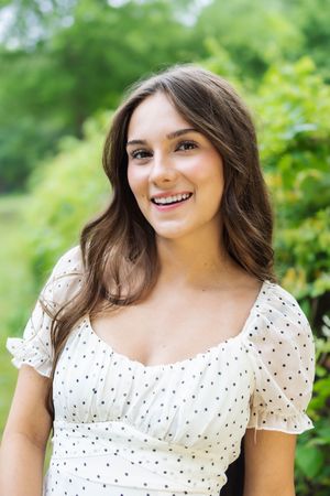 A portrait outdoors of someone wearing a white polka dot dress with puff sleeves against a green natural background.