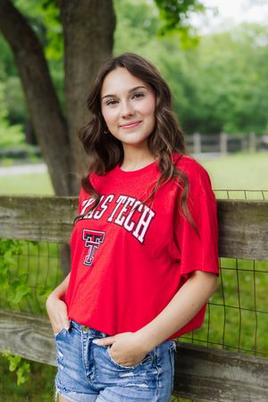 Student wearing a red Texas Tech t-shirt and denim shorts poses by rustic fence in outdoor park setting.