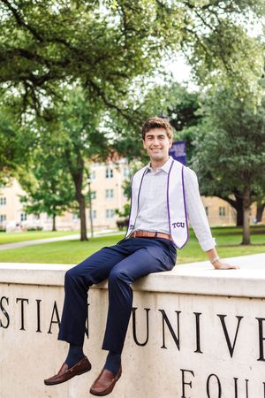 Someone wearing business casual attire with purple suspenders sits casually on a university sign wall in a leafy campus setting.