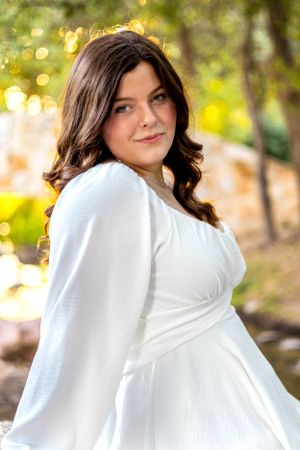 Portrait in a flowing white dress with long dark wavy hair against a sunlit natural outdoor background with bokeh.