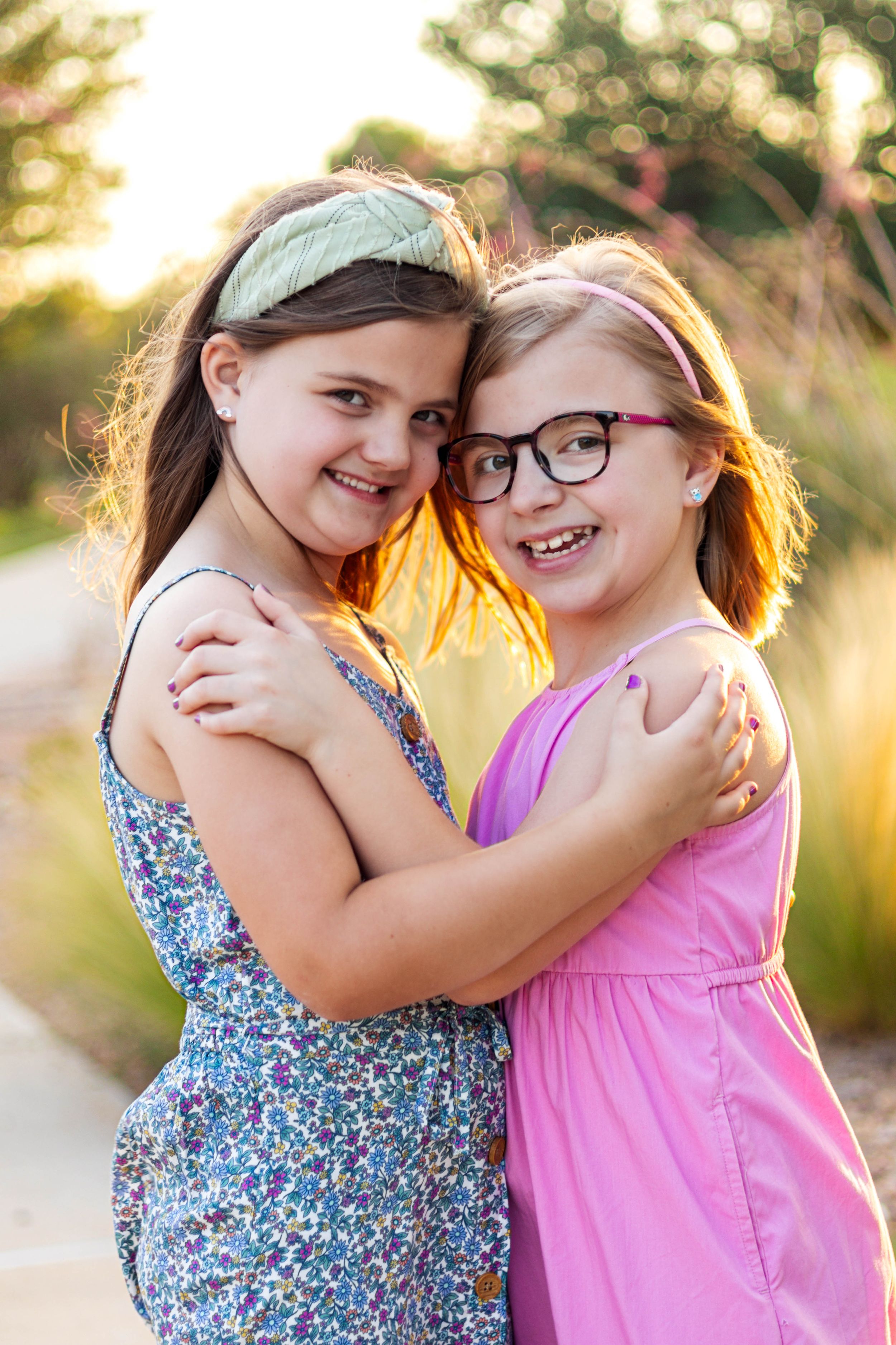Two young friends in summer dresses share a warm hug while smiling outdoors in golden sunlight.