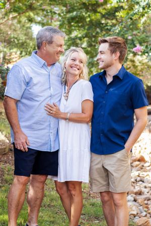 Three people share a candid moment outdoors smiling together in casual summer attire against a natural backdrop.
