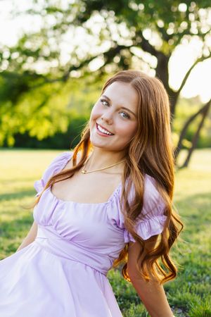 A young woman in a lilac summer dress poses outdoors in a sunny park with trees and natural lighting.