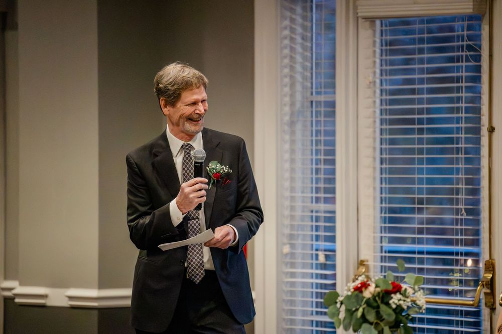 A groom in a black suit stands in front of windows at a wedding ceremony venue with floral decorations.