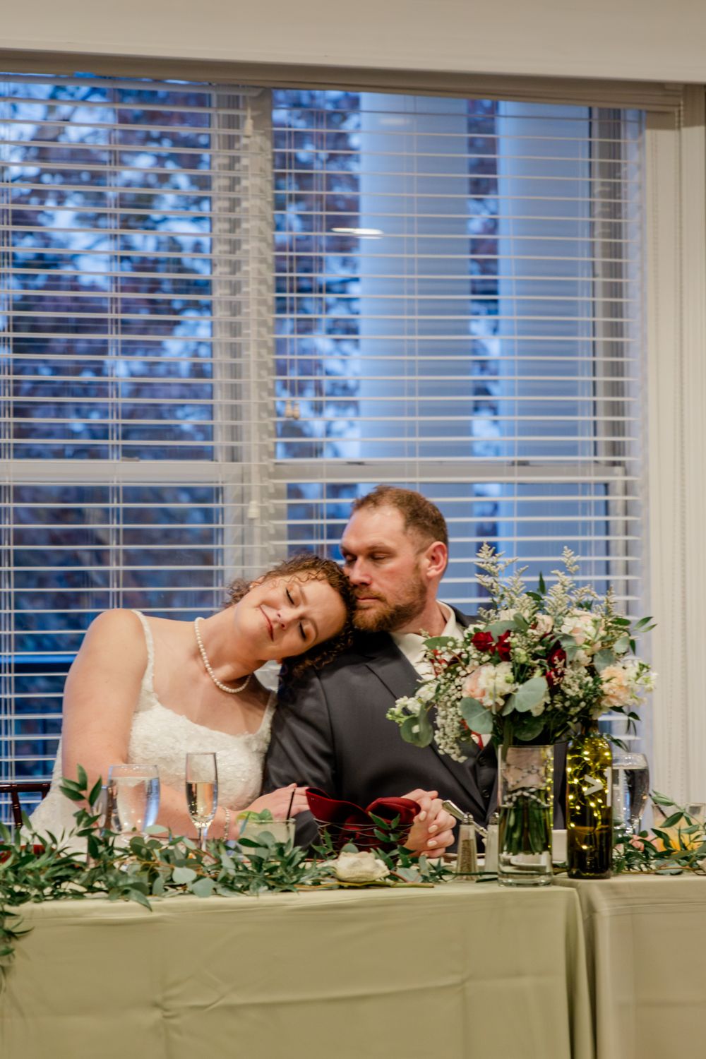 A romantic couple shares an intimate moment at their wedding reception table decorated with greenery and floral arrangements.