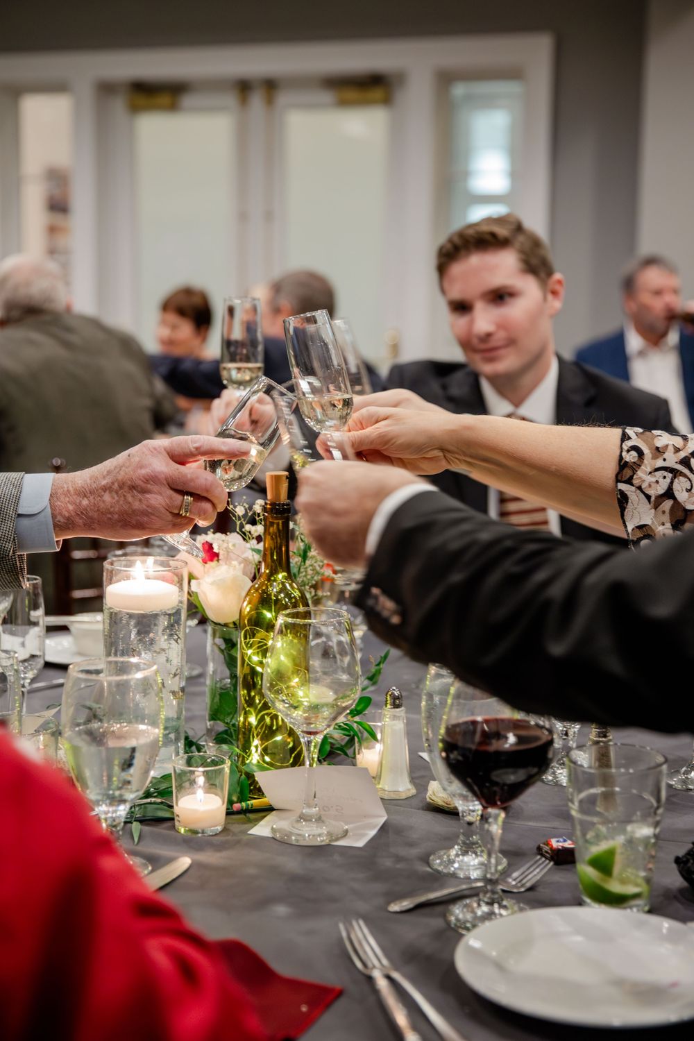 Hands reach across an elegant dinner table set with wine glasses, candles, and bottles during a social gathering.