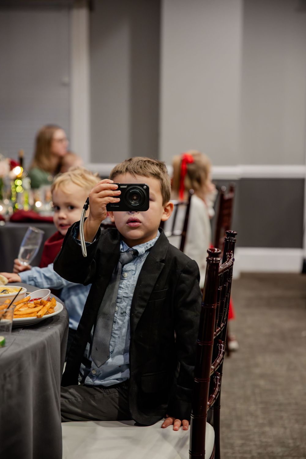 A young child in a suit jacket sits at a dining table during a formal event.