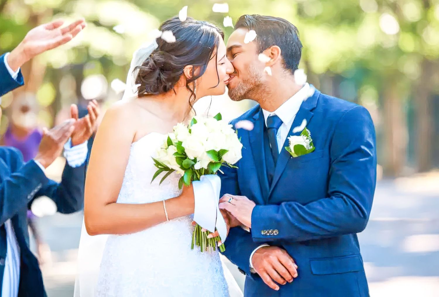 A bride in white dress and groom in navy suit share an intimate moment while holding a white floral bouquet outdoors.