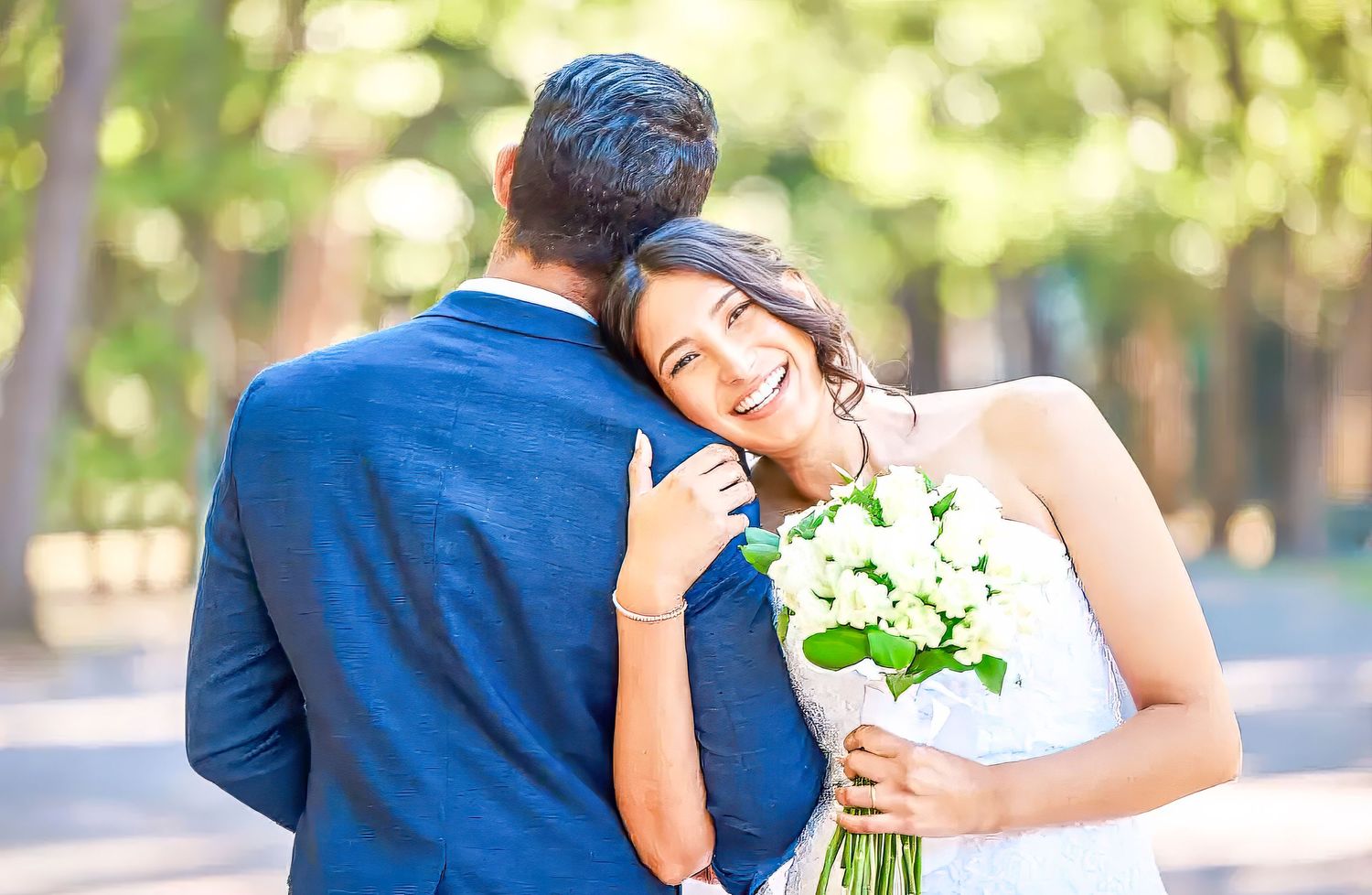 A romantic wedding couple embraces outdoors in a garden setting with soft natural lighting and white floral bouquet.