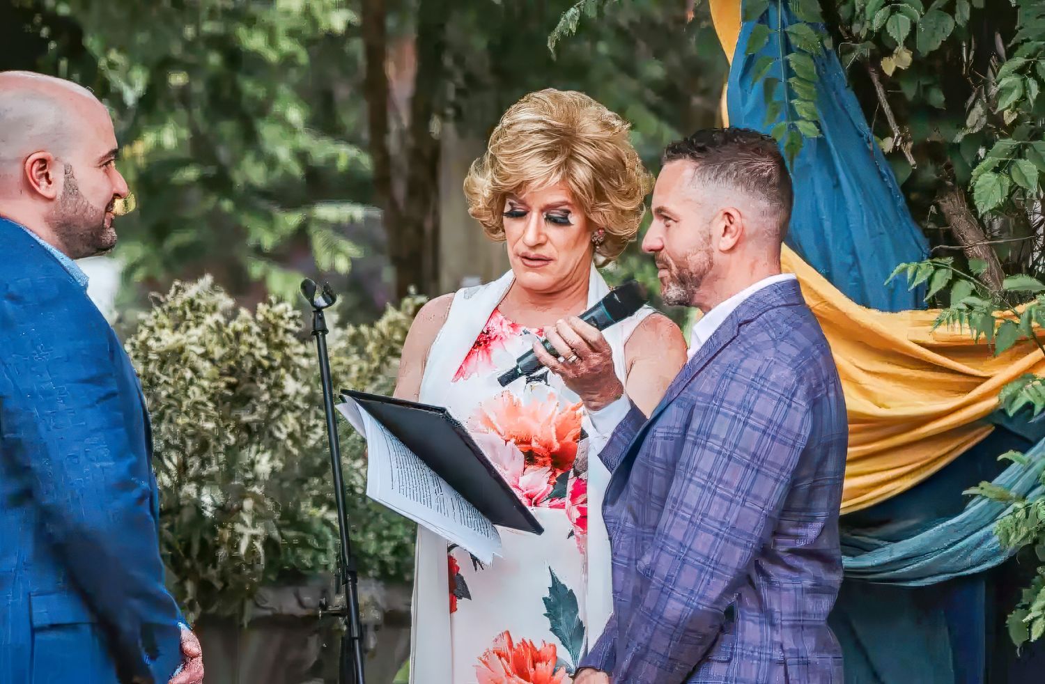 A wedding ceremony takes place outdoors with gold draping and green foliage in the background.
