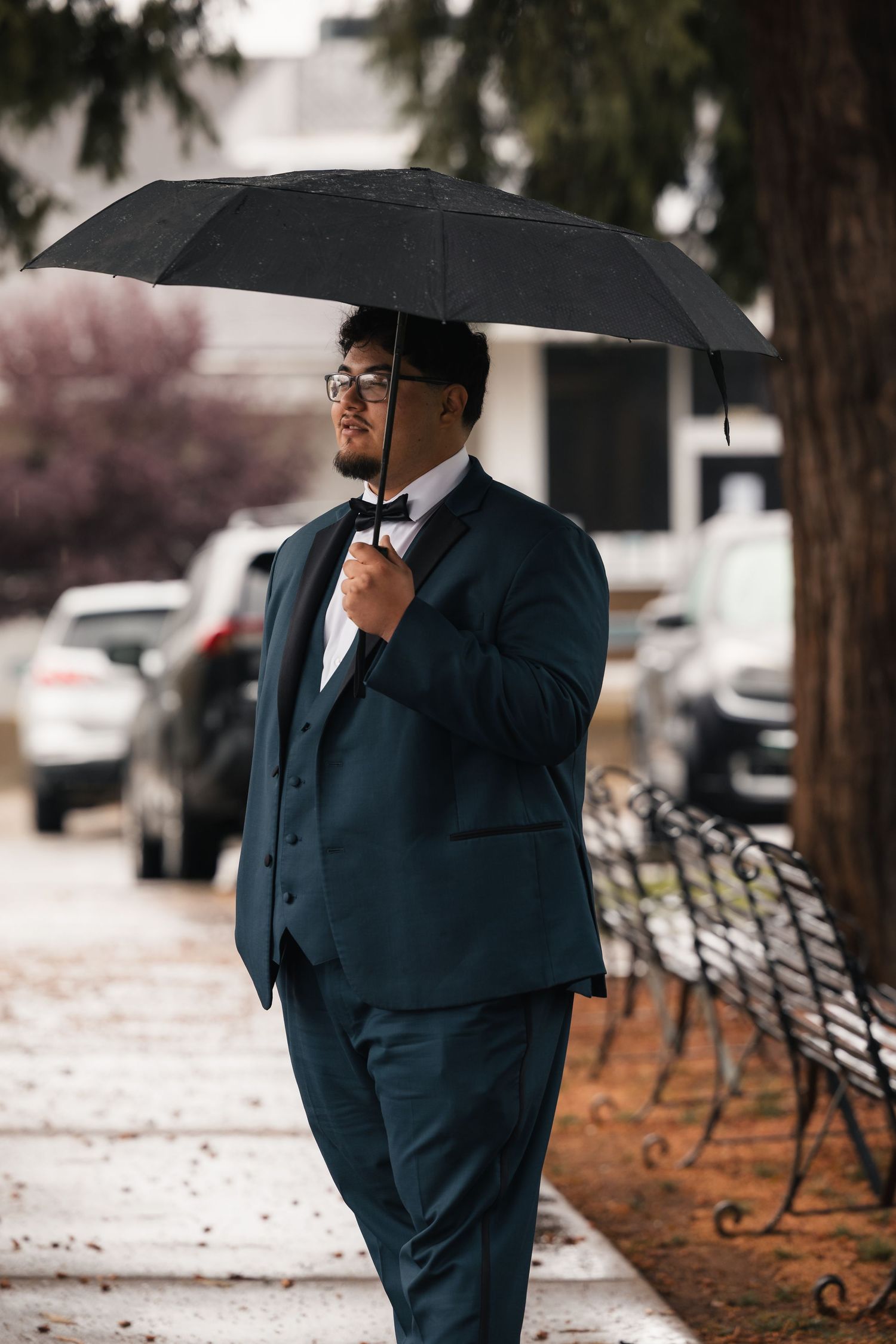 A groom in a navy suit and bow tie stands with a black umbrella on a rainy day.
