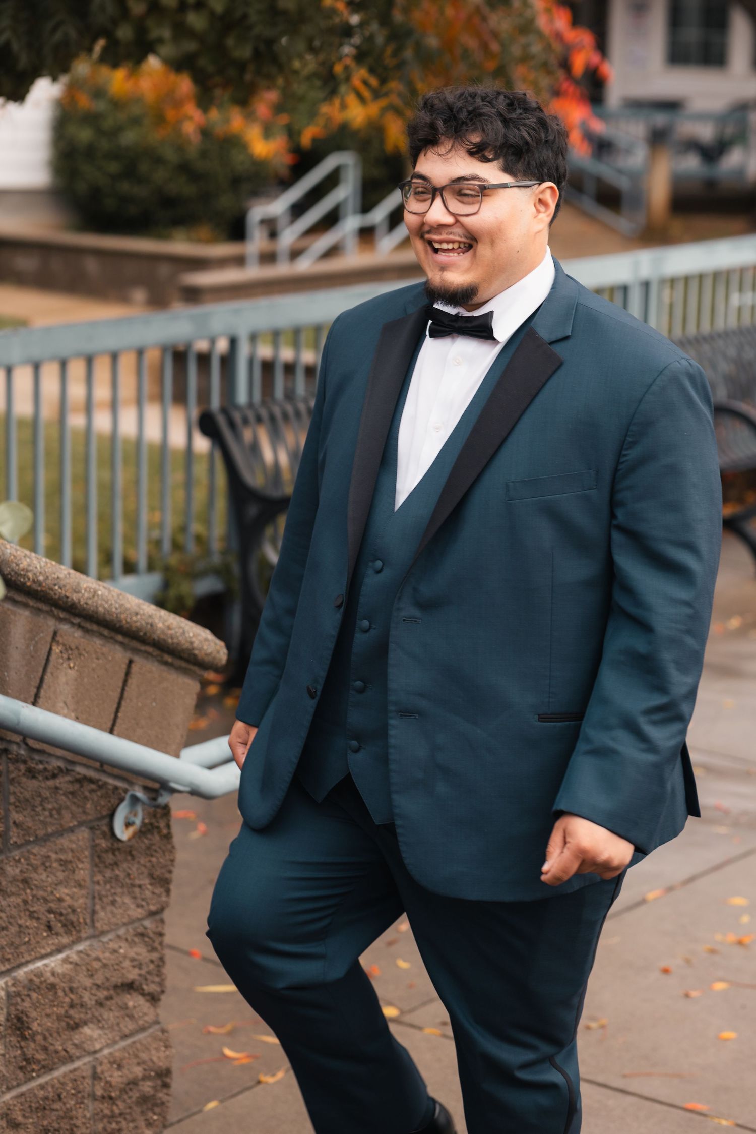 A person in a navy suit and white shirt smiles while standing near an outdoor railing on an autumn day.