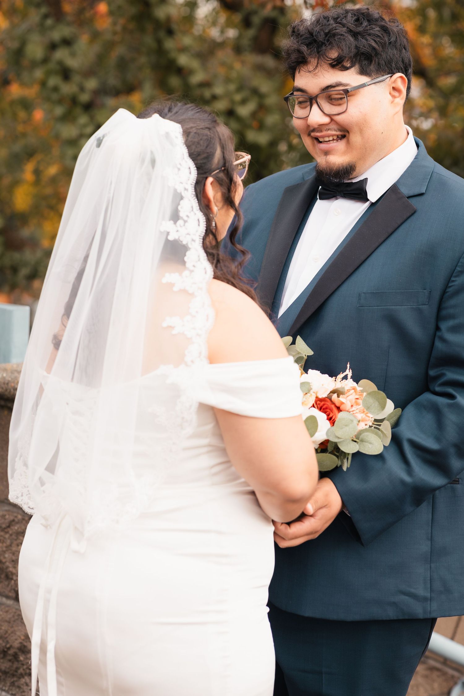 A romantic wedding moment between a bride in white dress with veil and groom in navy suit during an autumn day.