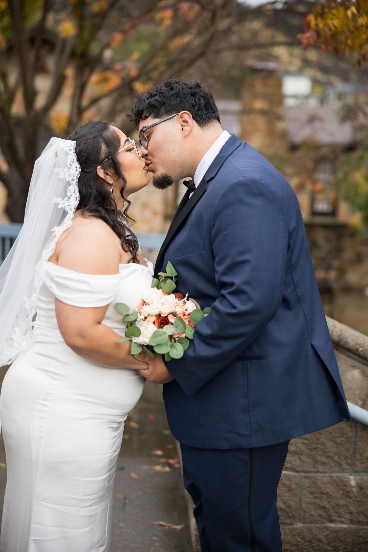 A romantic wedding couple shares a kiss outdoors during autumn with fall foliage in the background.
