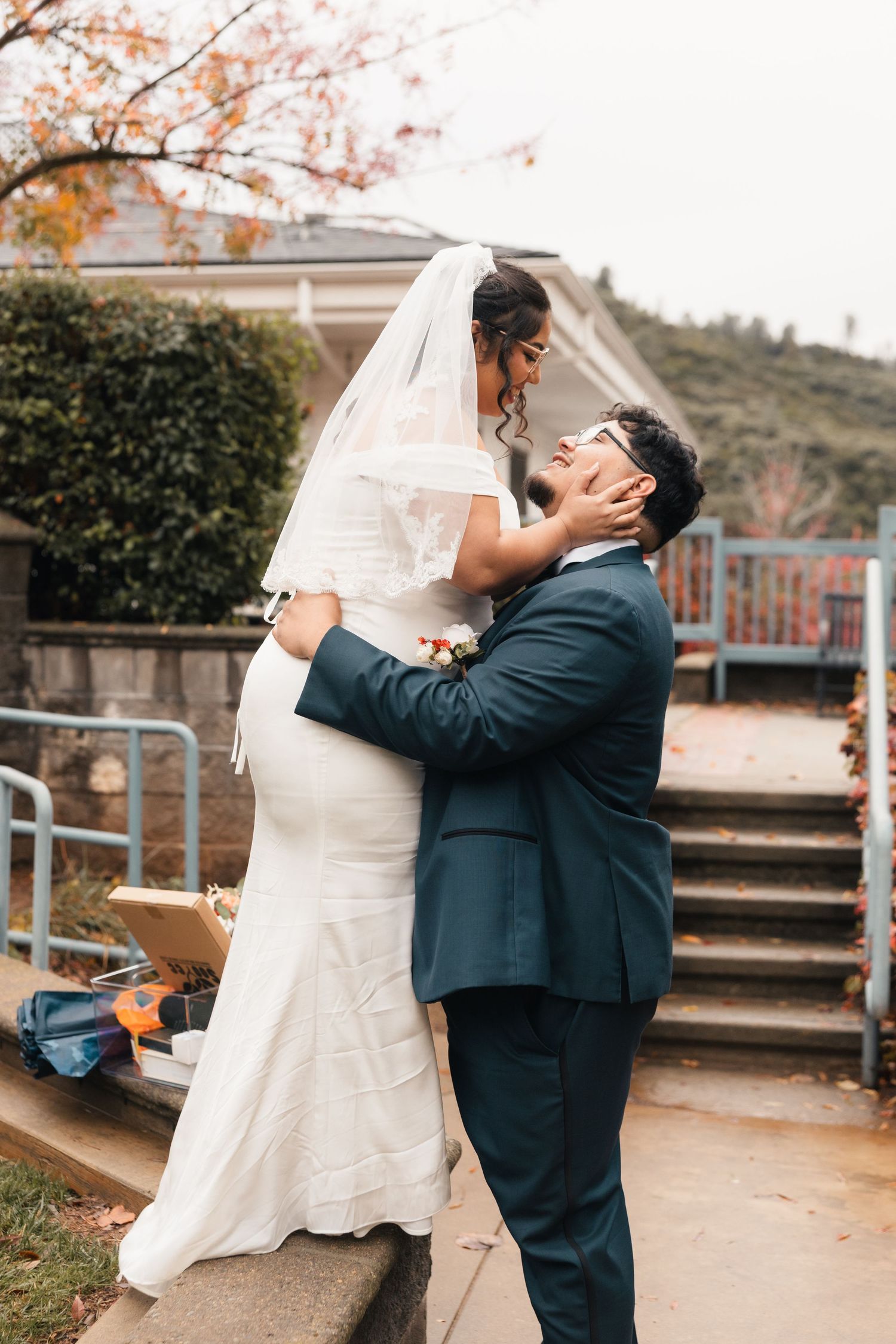 Two people in formal wedding attire embrace joyfully outdoors with autumn foliage in the background.