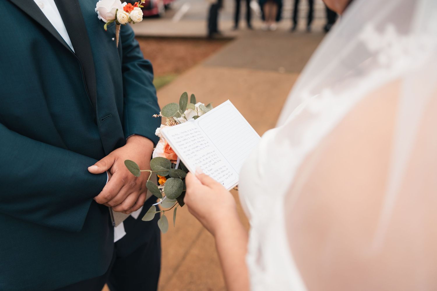 Close-up of hands exchanging wedding vows during outdoor ceremony with white veil and dark suit visible.