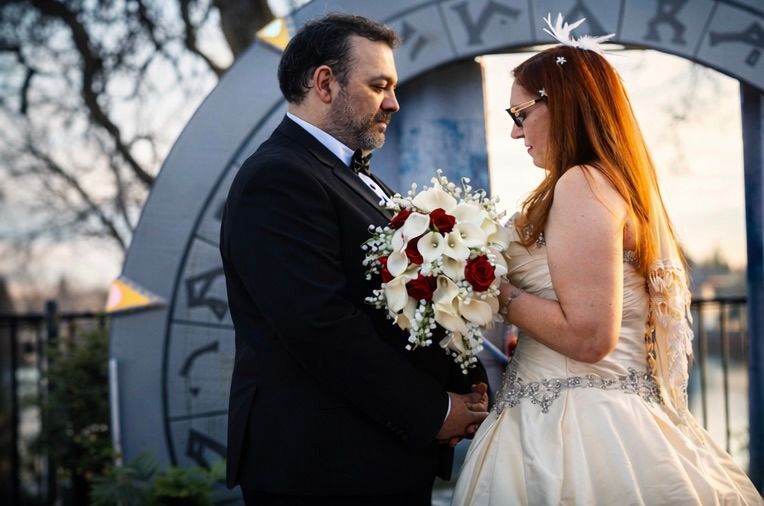 Bride and groom holding red and white bouquet stand face to face before zodiac arch at outdoor wedding ceremony.