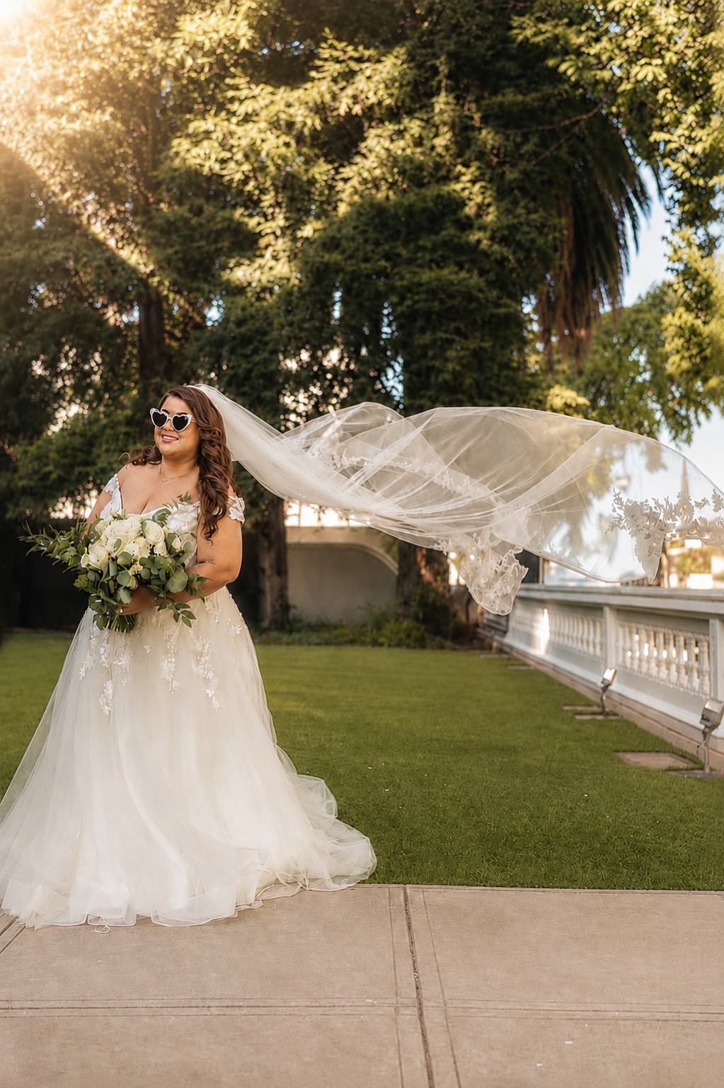 Bride in white ball gown with flowing veil and sunglasses holding bouquet outdoors in golden sunlight.