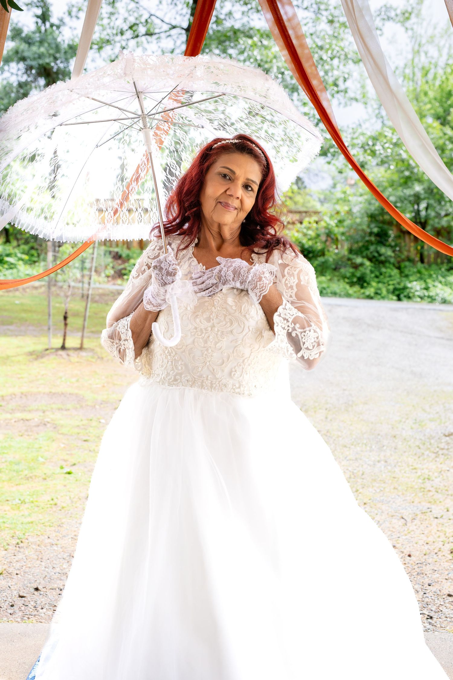 A bride in a white lace gown holds a decorative umbrella outdoors under colorful ribbons.