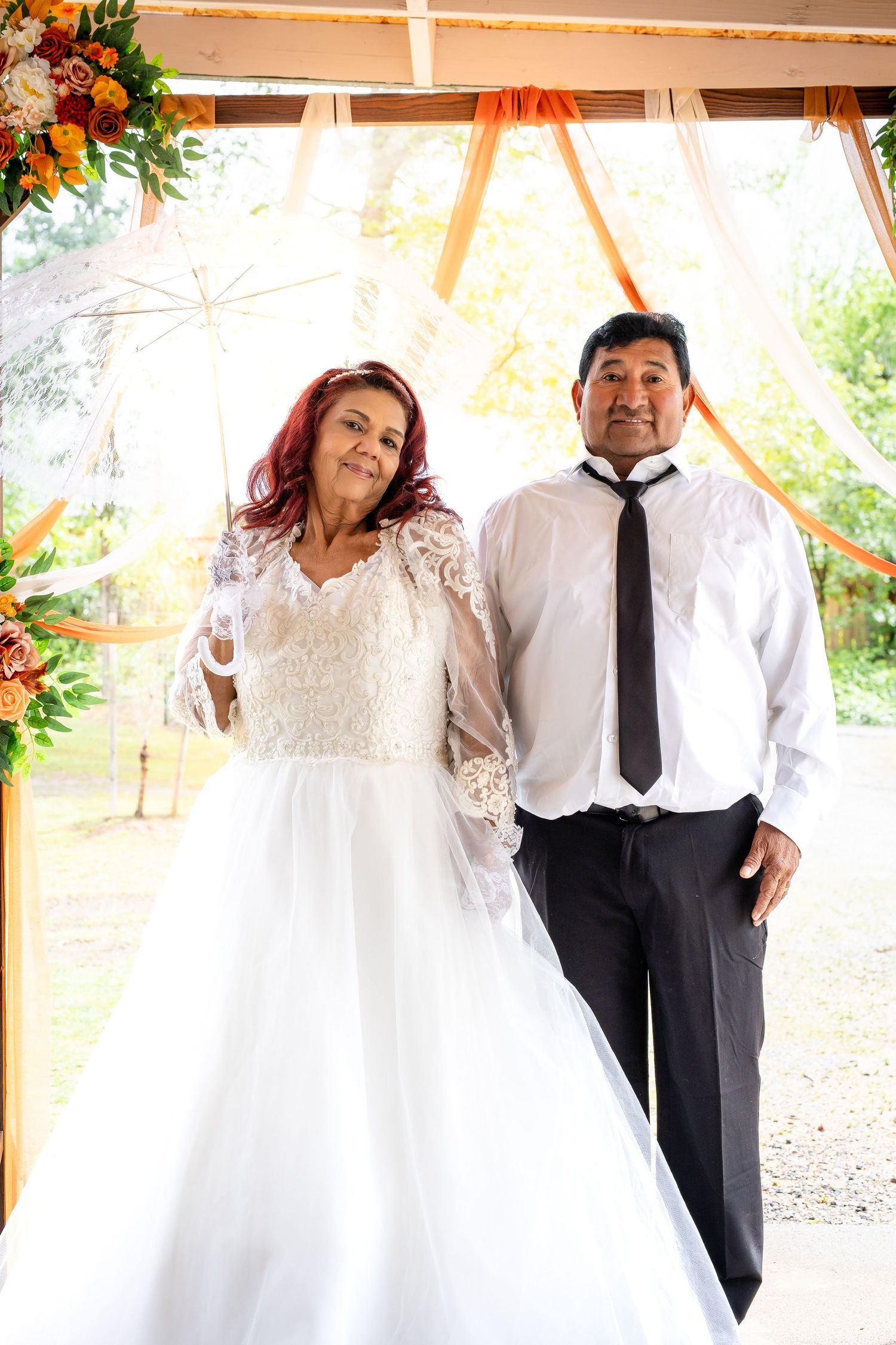 Happy bride in white lace gown and groom in black tie posing under floral wedding arch.