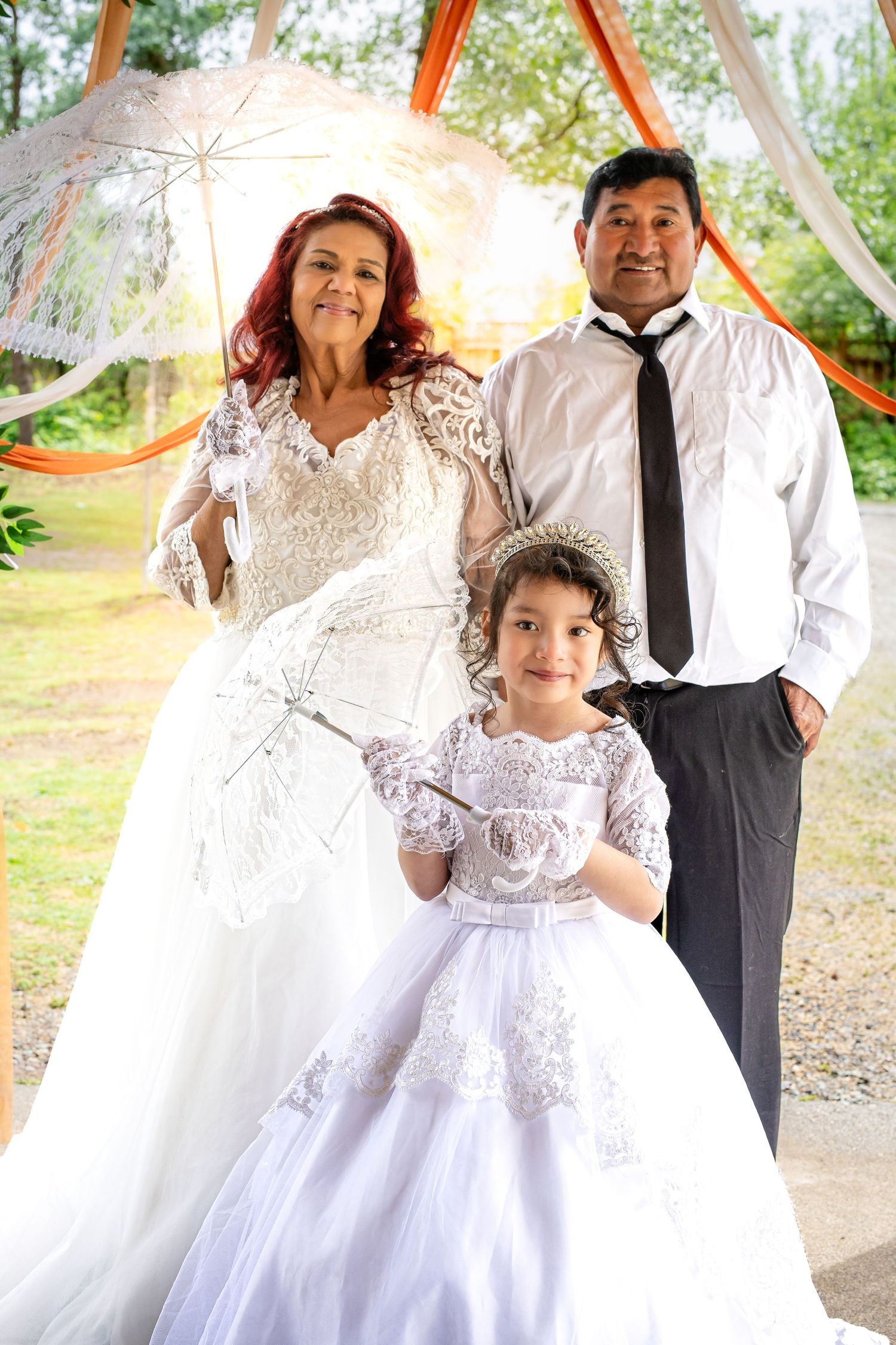 Bride and groom pose with young flower girl in white dress holding lace umbrella at outdoor wedding ceremony.