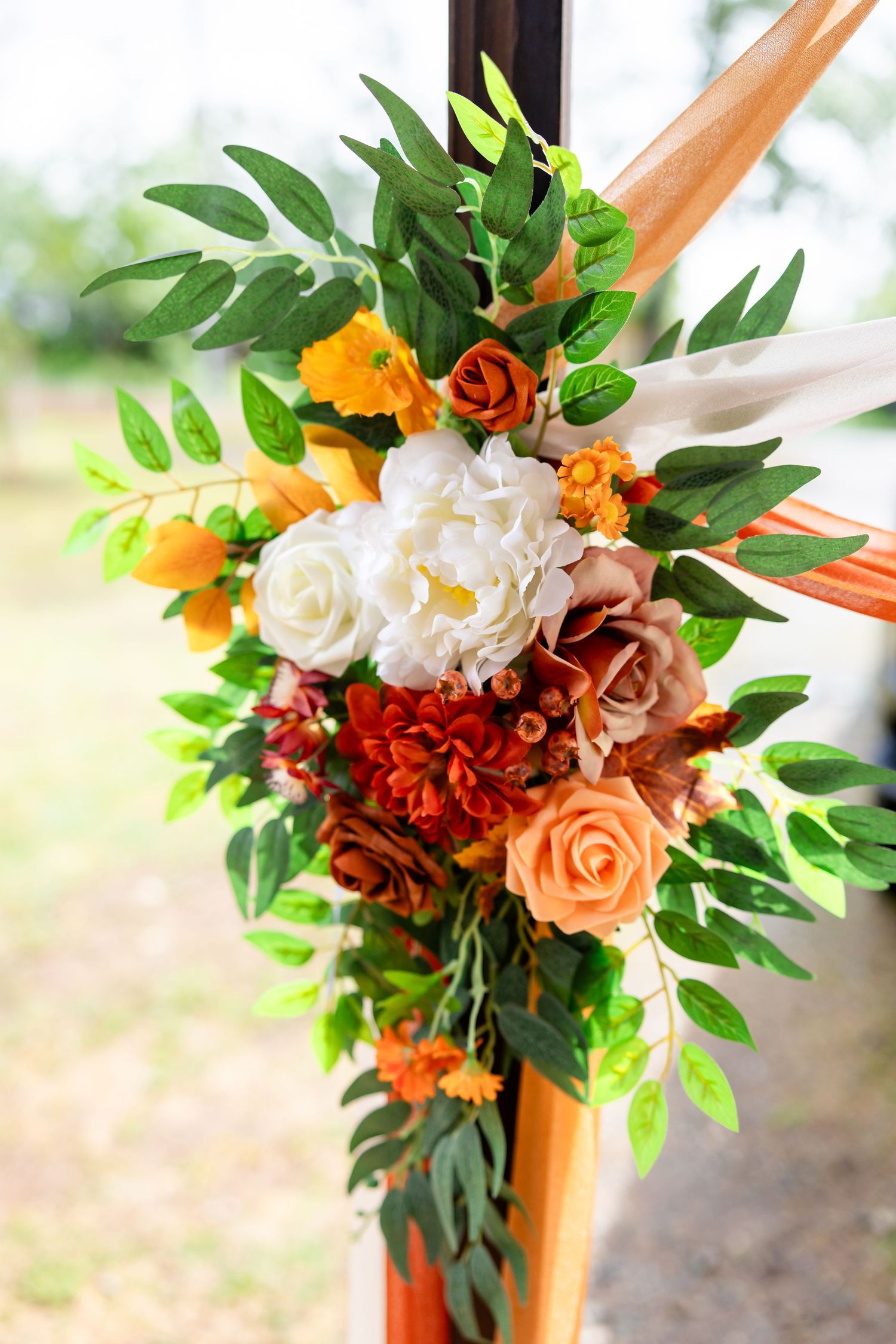 Colorful wedding floral arrangement with white, orange, and red roses, greenery, and ribbon on a wooden arch post.