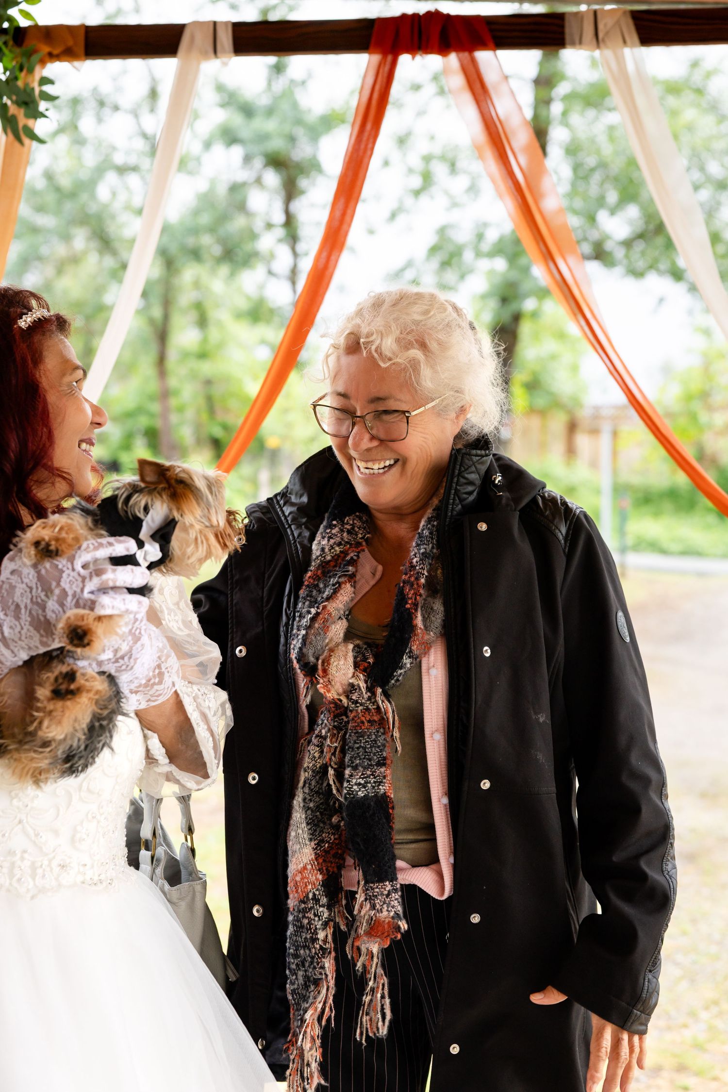 Bride holding a Yorkshire Terrier laughs with an older woman in glasses at an outdoor wedding ceremony.