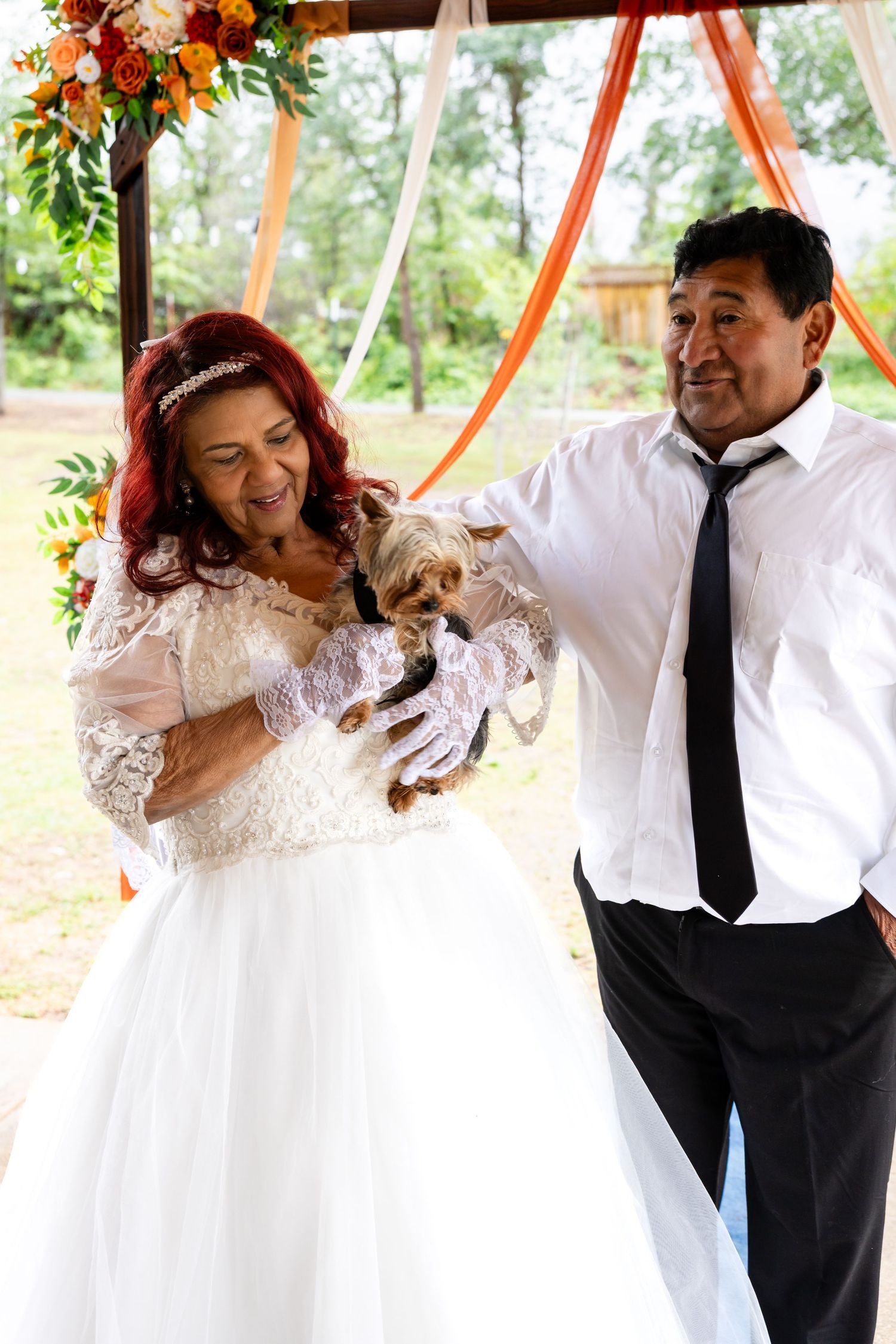 Smiling bride in lace gown holds small Yorkshire Terrier dog beside groom at outdoor wedding celebration.