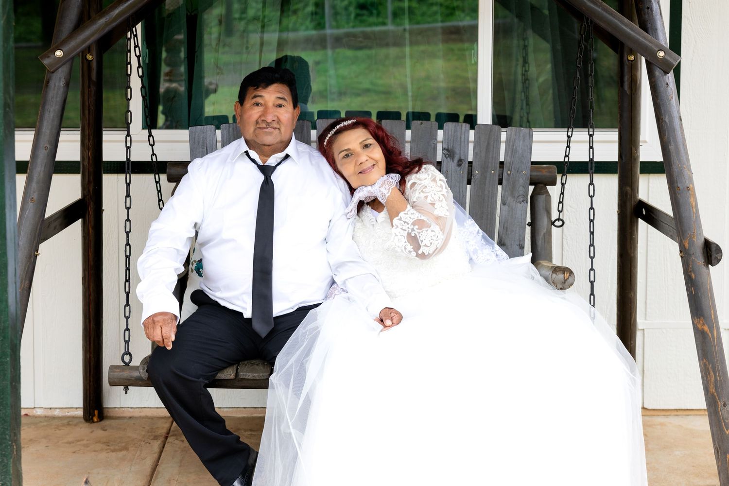 Bride and groom sit together on wooden porch swing in wedding attire for romantic portrait photo.