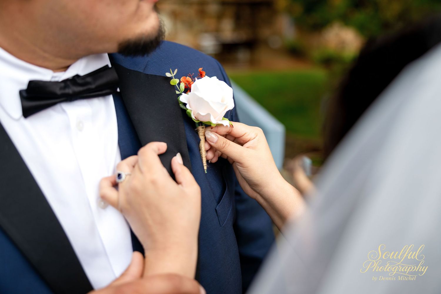 A white rose boutonniere is being pinned onto a navy blue suit jacket at a formal wedding ceremony.