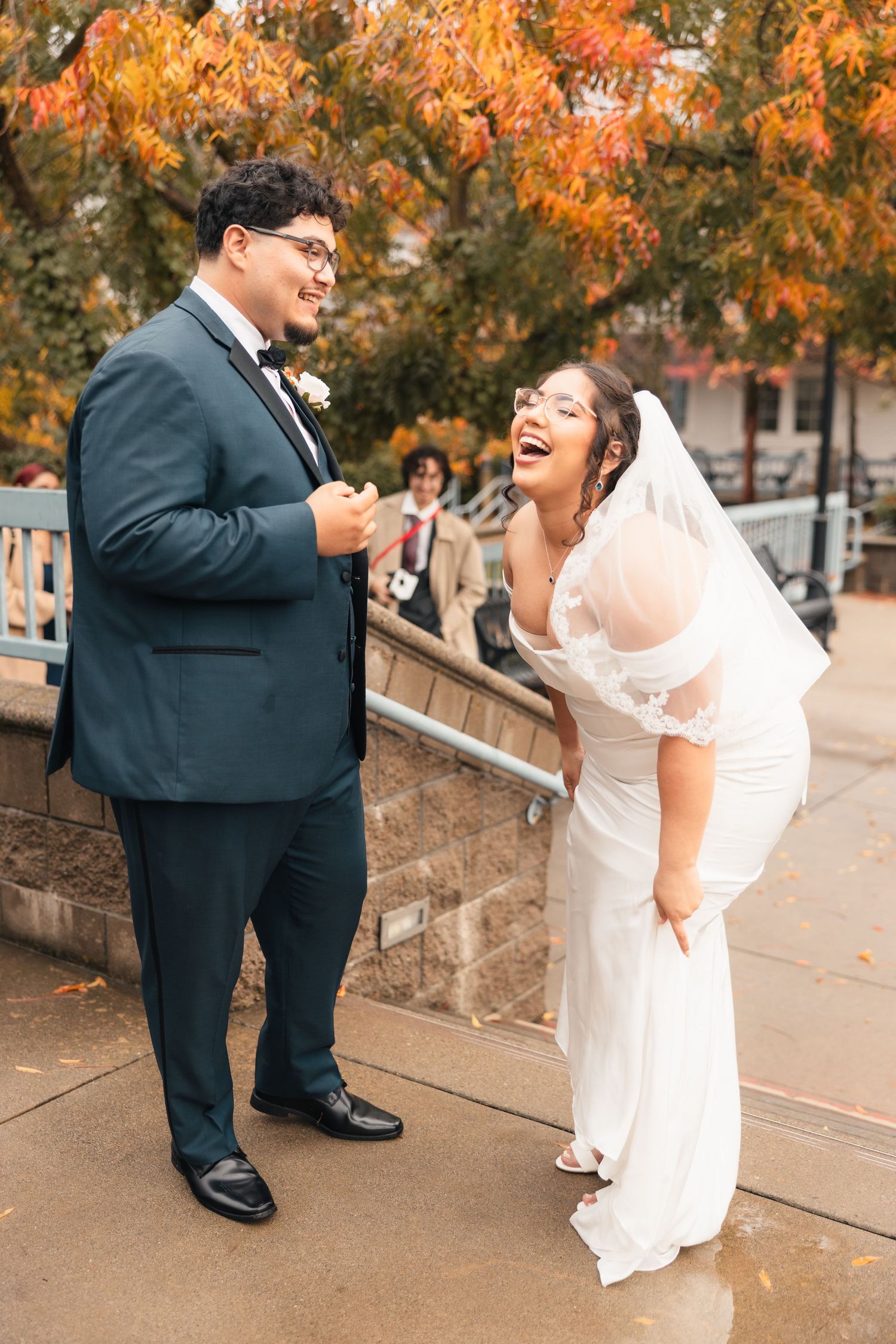 A couple shares a joyful moment together on stone steps surrounded by autumn foliage in their wedding attire.