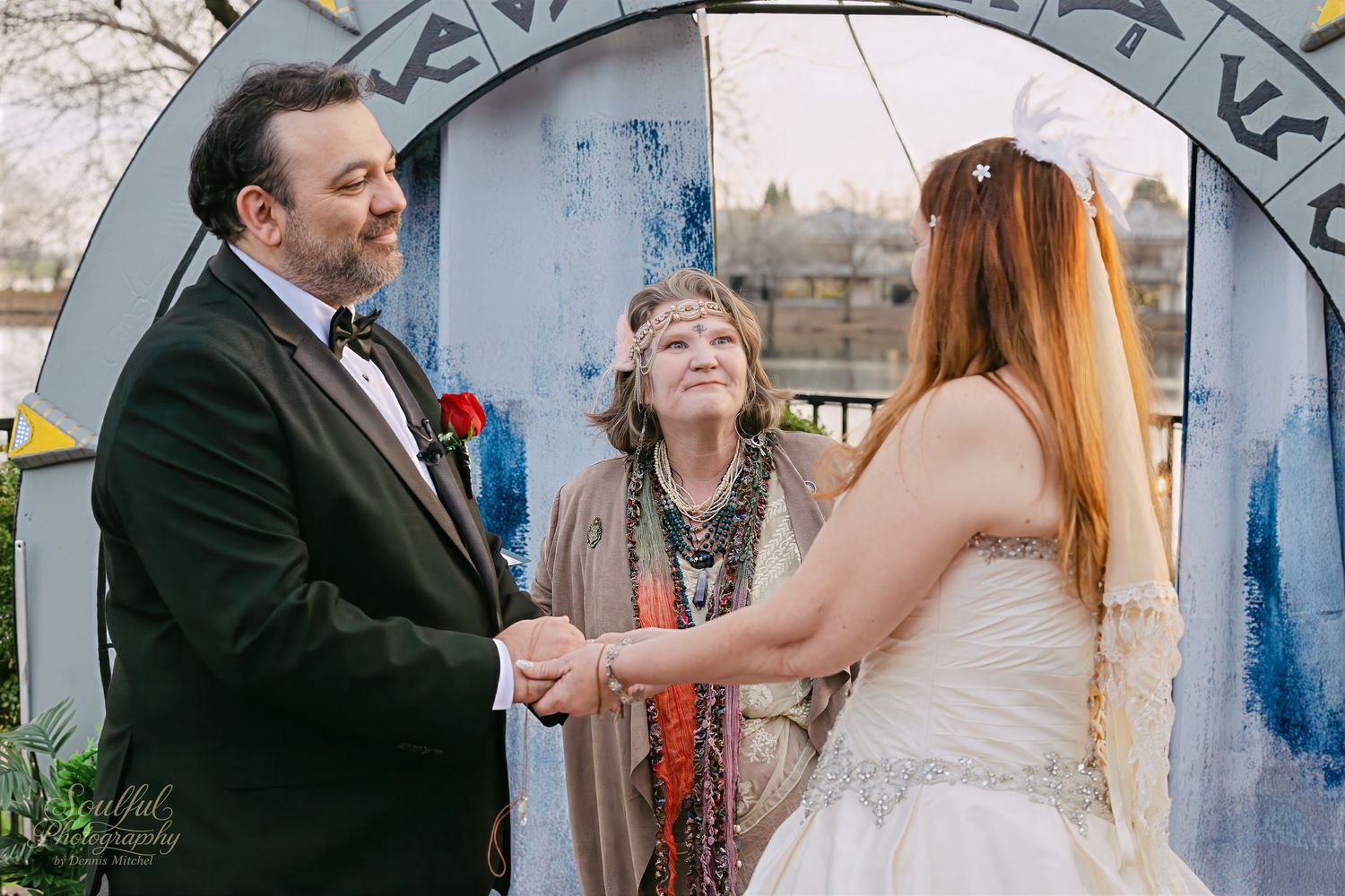 A couple shares a joyful moment with an officiant during their outdoor wedding ceremony under a blue archway.