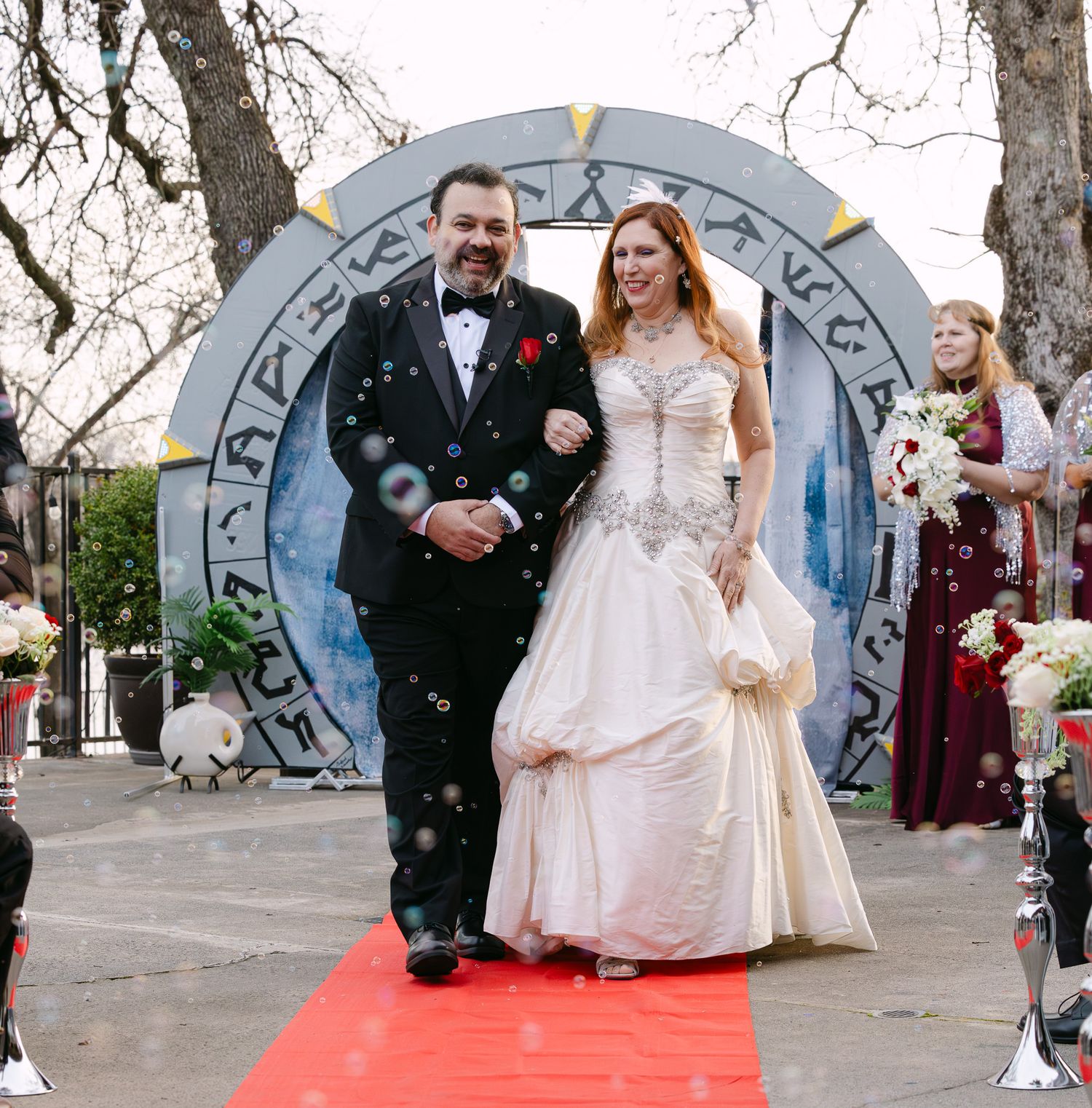 Wedding couple walks down a red carpet path outdoors with guests throwing confetti in a winter garden setting.