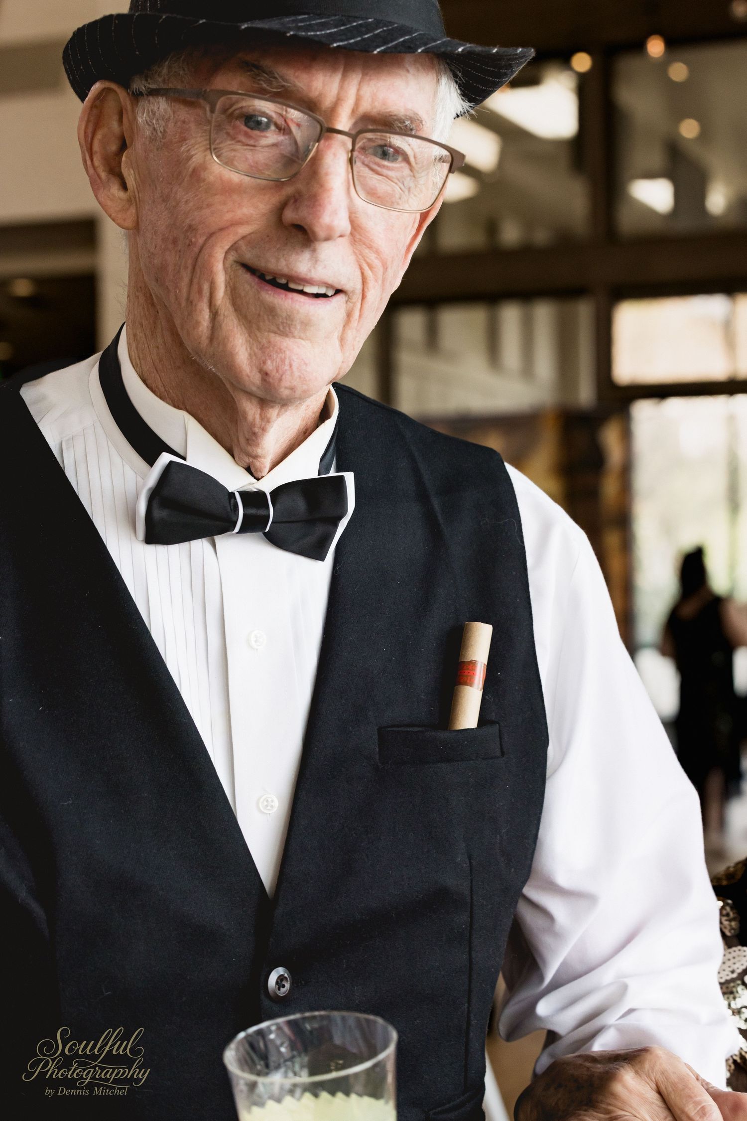 Elderly man in traditional black vest and white shirt smiles warmly while serving at a restaurant.