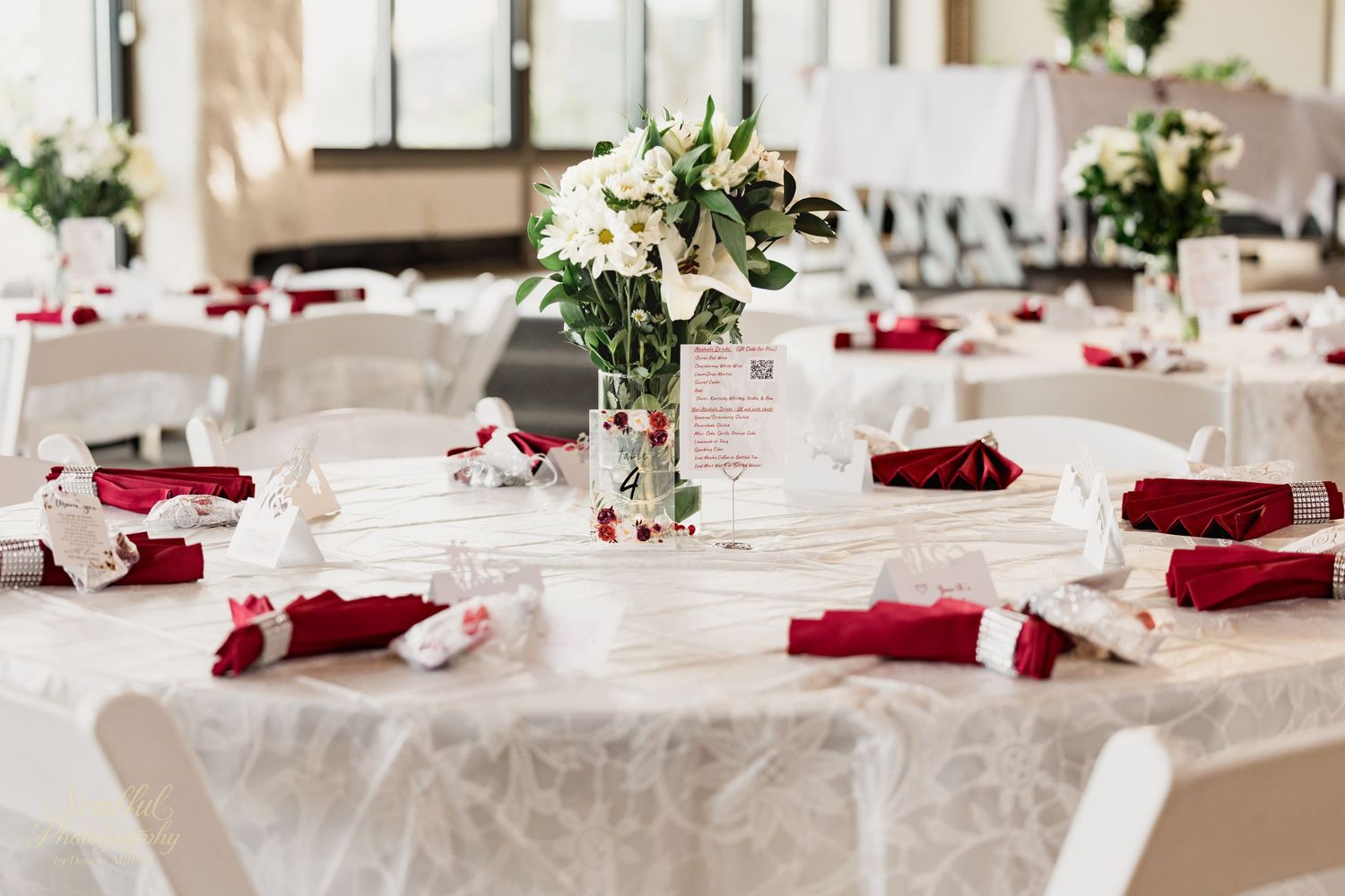 Elegant wedding reception tables decorated with red napkins, white linens and greenery centerpieces in a bright venue.