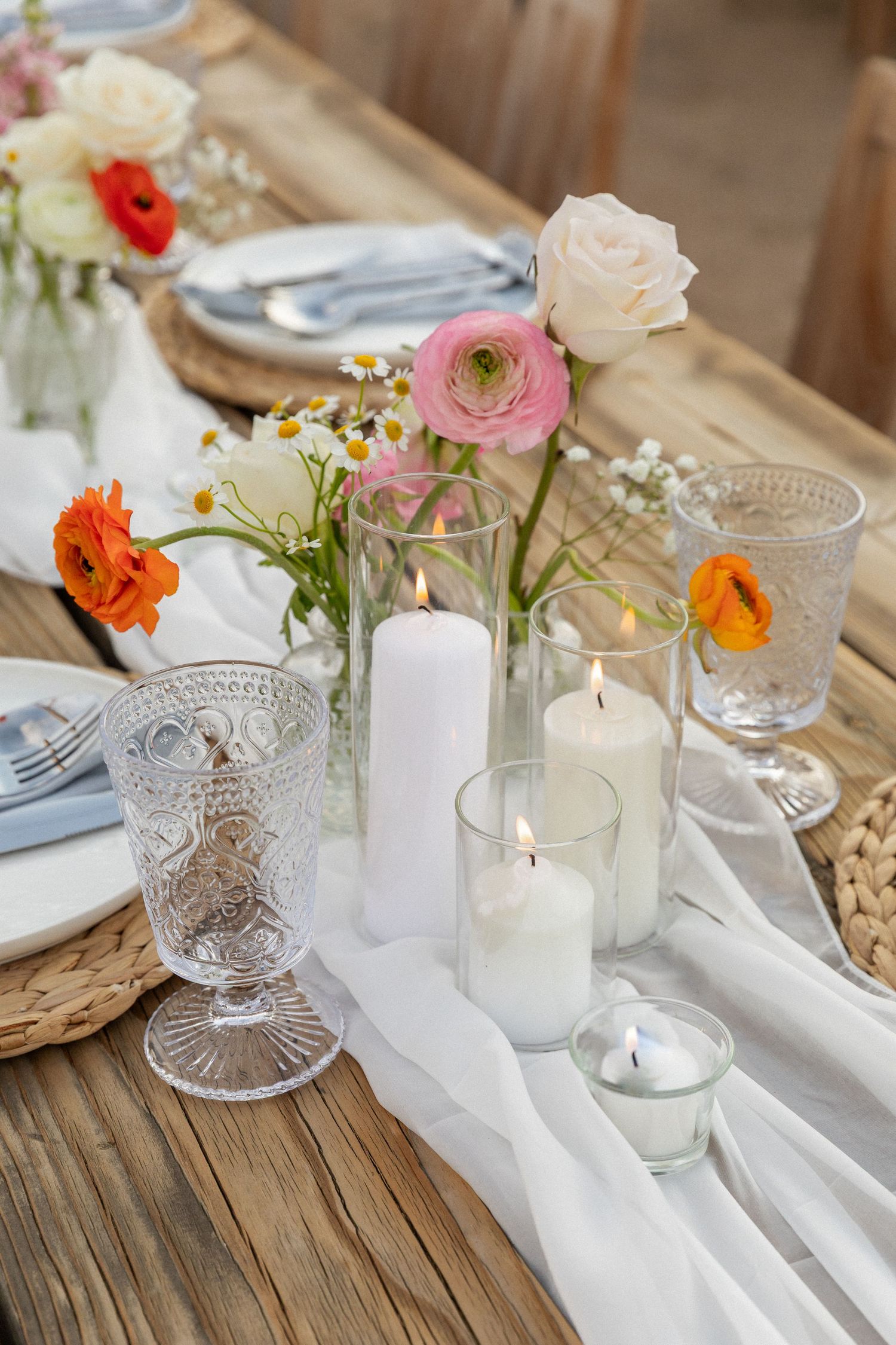 Elegant table setting with white candles, pink ranunculus flowers, and vintage crystal glasses on a wooden surface.
