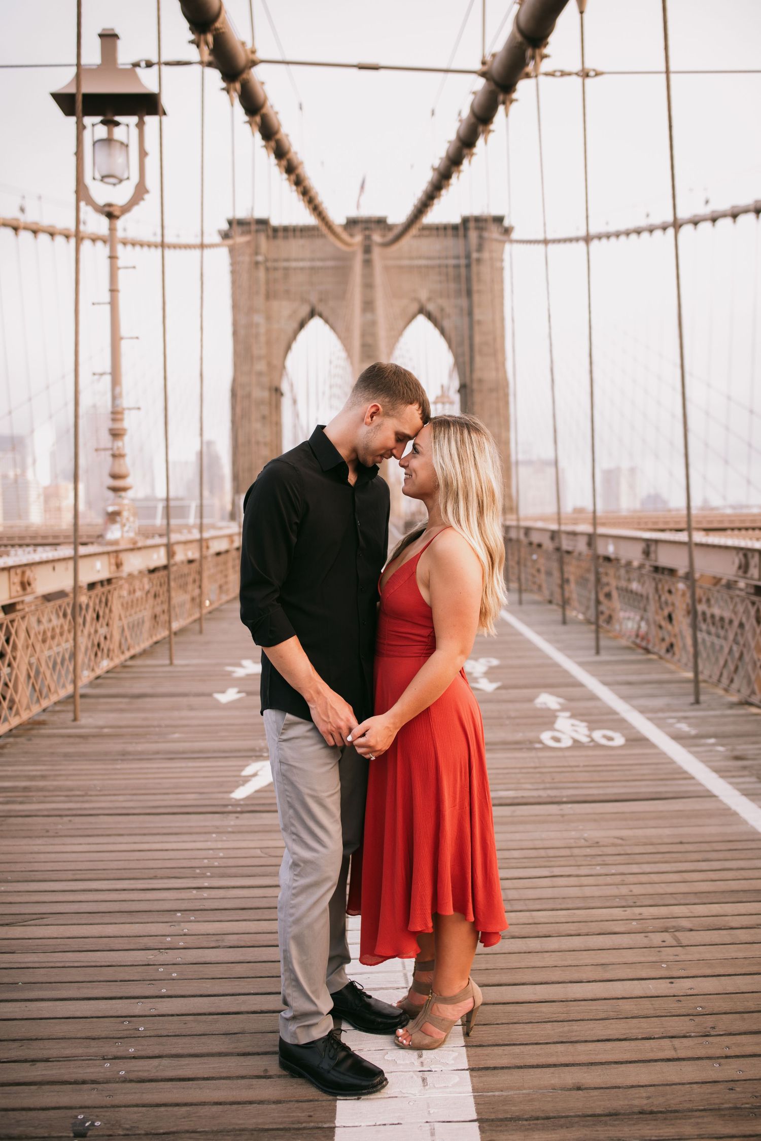 Romantic couple in red dress and black shirt embrace on the Brooklyn Bridge at golden hour.