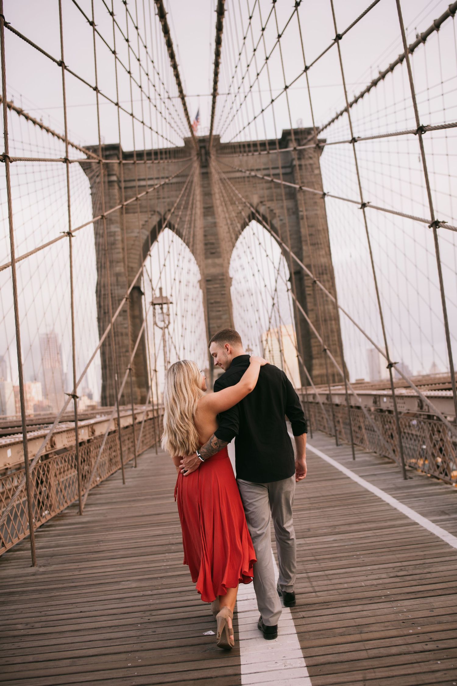 Romantic couple in red dress and black shirt embrace on the Brooklyn Bridge at golden hour.