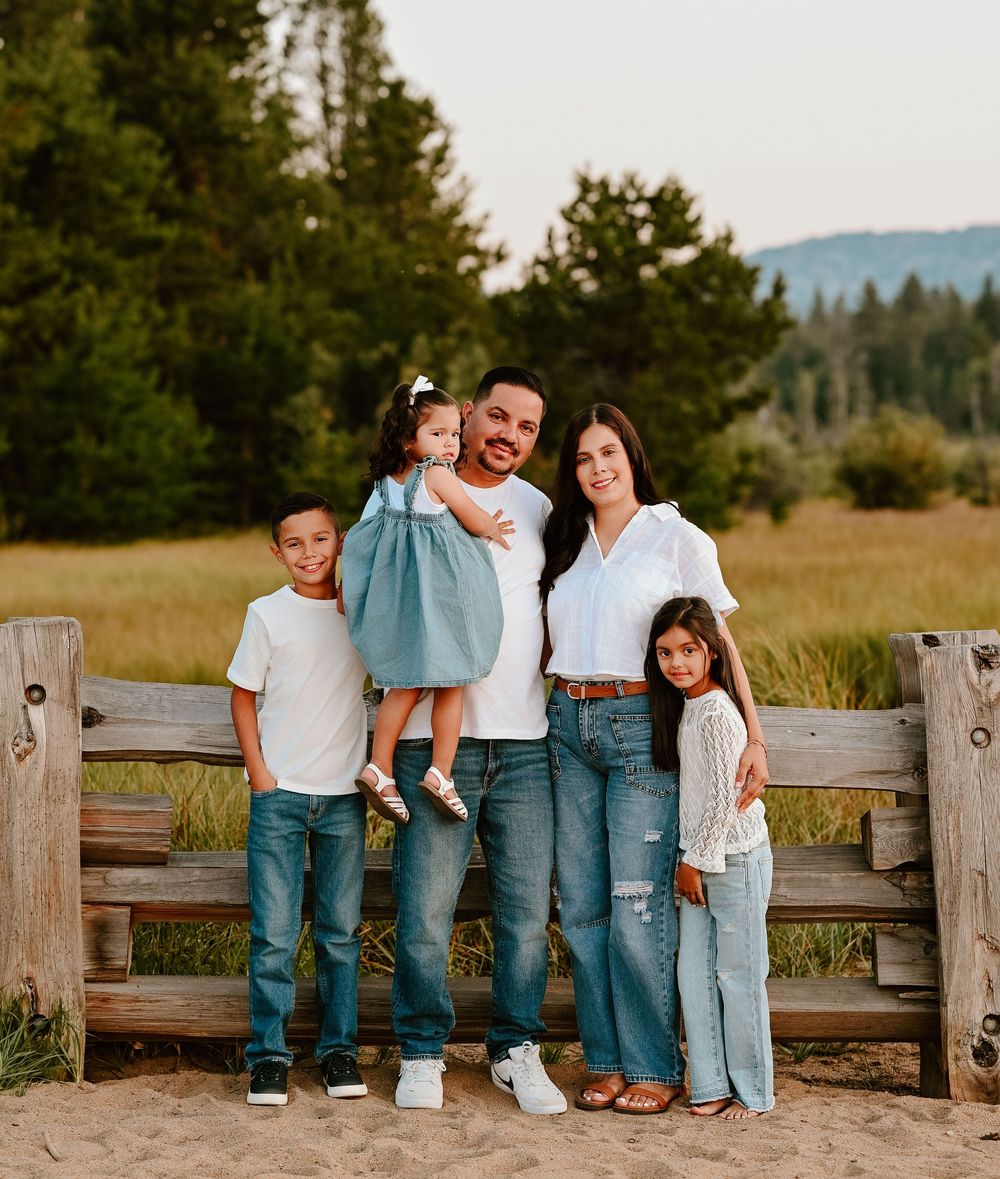 A family in coordinating denim outfits poses casually along a wooden fence in a rustic outdoor setting at sunset.