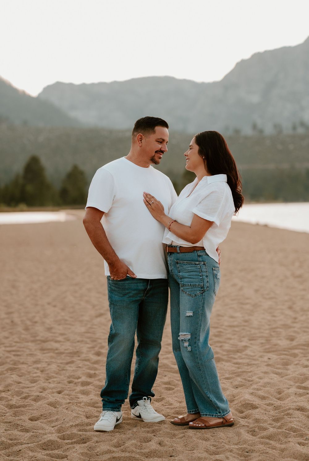 A couple shares a joyful moment together on a sandy beach with mountains in the background at sunset.