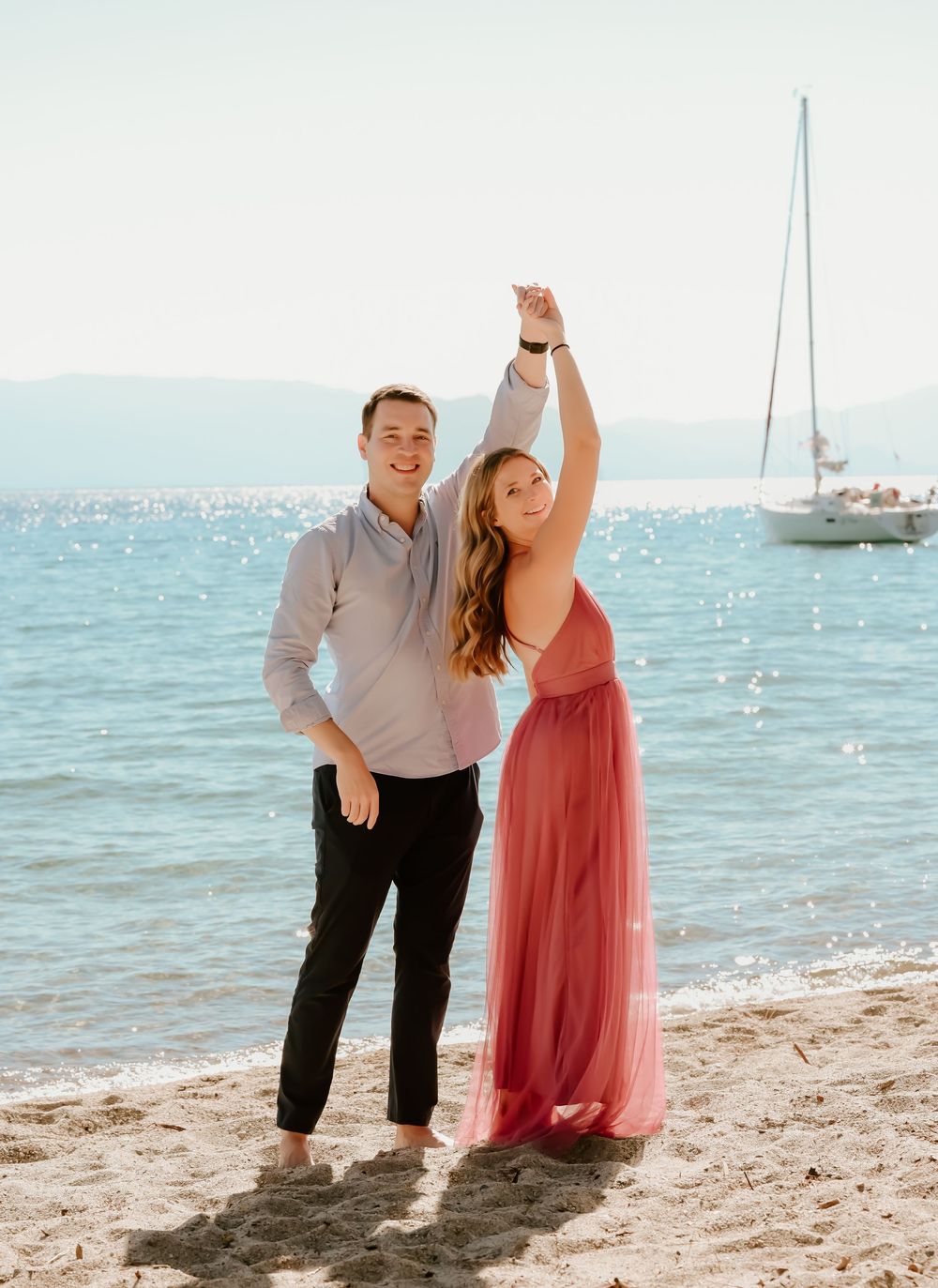 A couple dancing on a sunny beach with a sailboat in the background and sparkling blue water.
