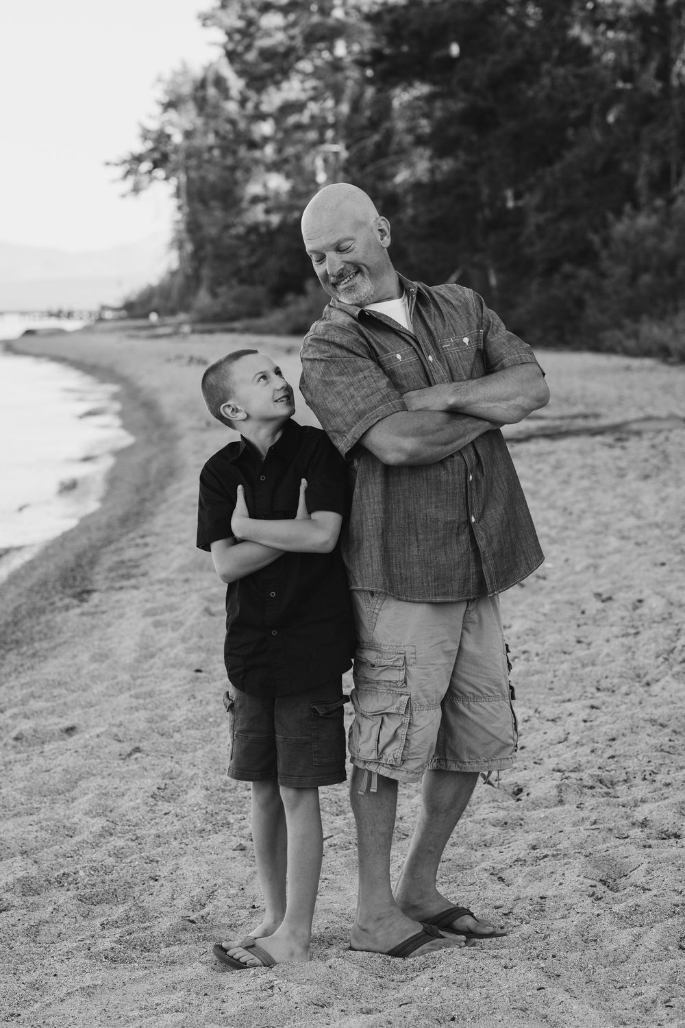 Black and white portrait of two people standing together on a sandy beach with trees in the background.